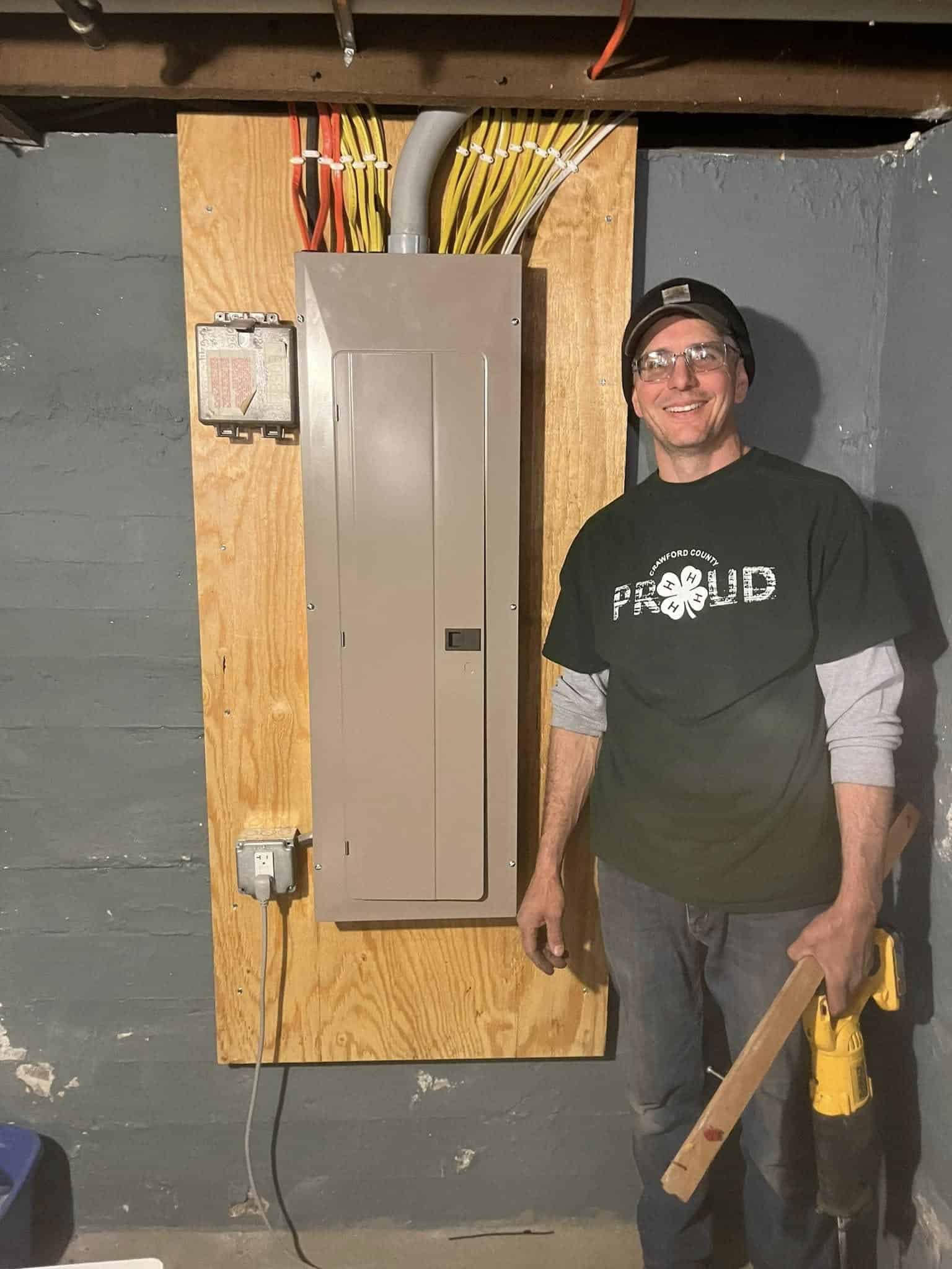 A smiling man stands next to an electrical panel mounted on a wooden board, holding a drill and a piece of wood in a basement with exposed walls and ceiling.