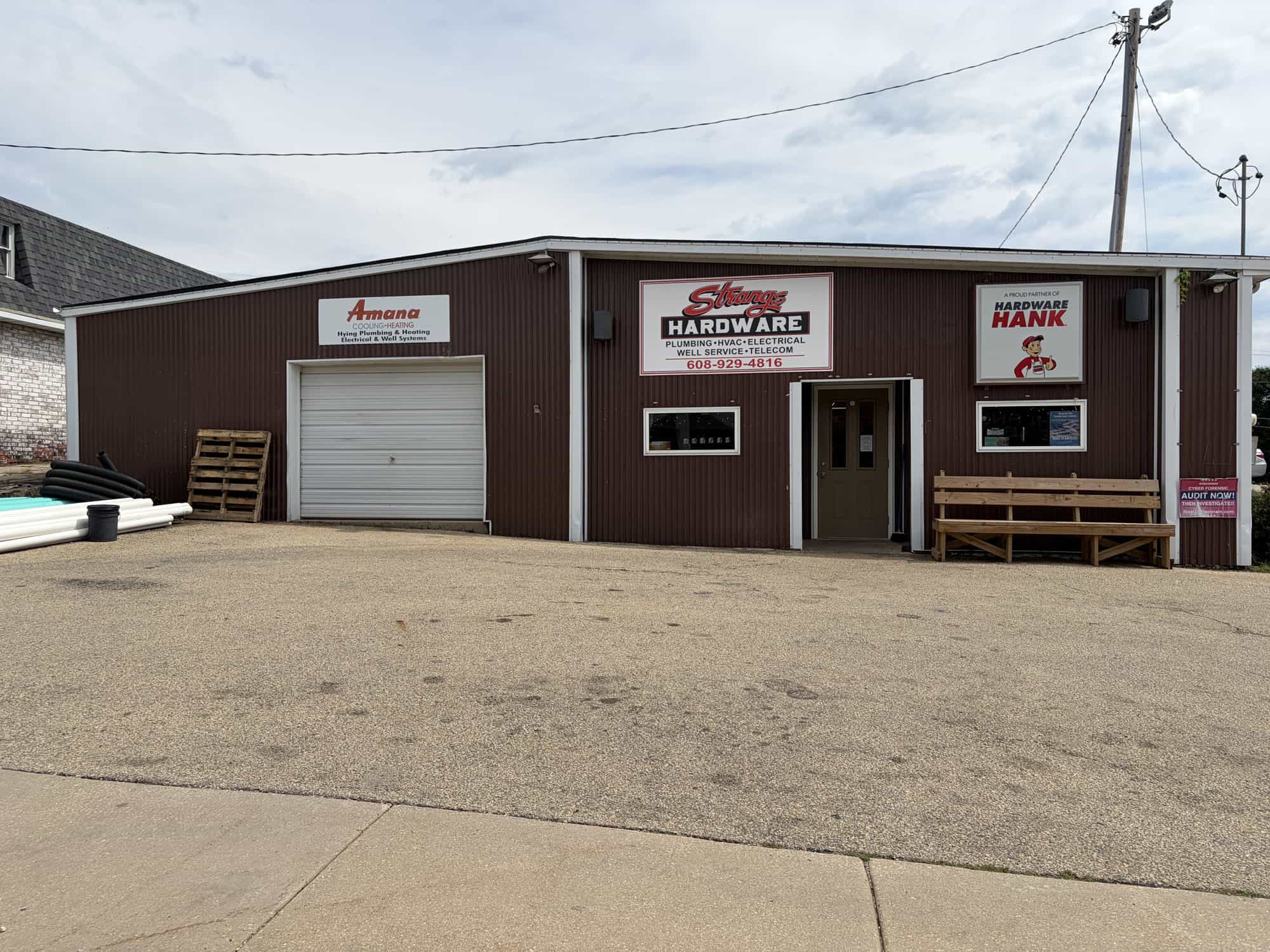 A brown hardware store with signs for Amana, Sherry Hardware, and Hardware Hank. There is a garage door, a regular door, benches, and some pipes and pallets outside on a paved area.