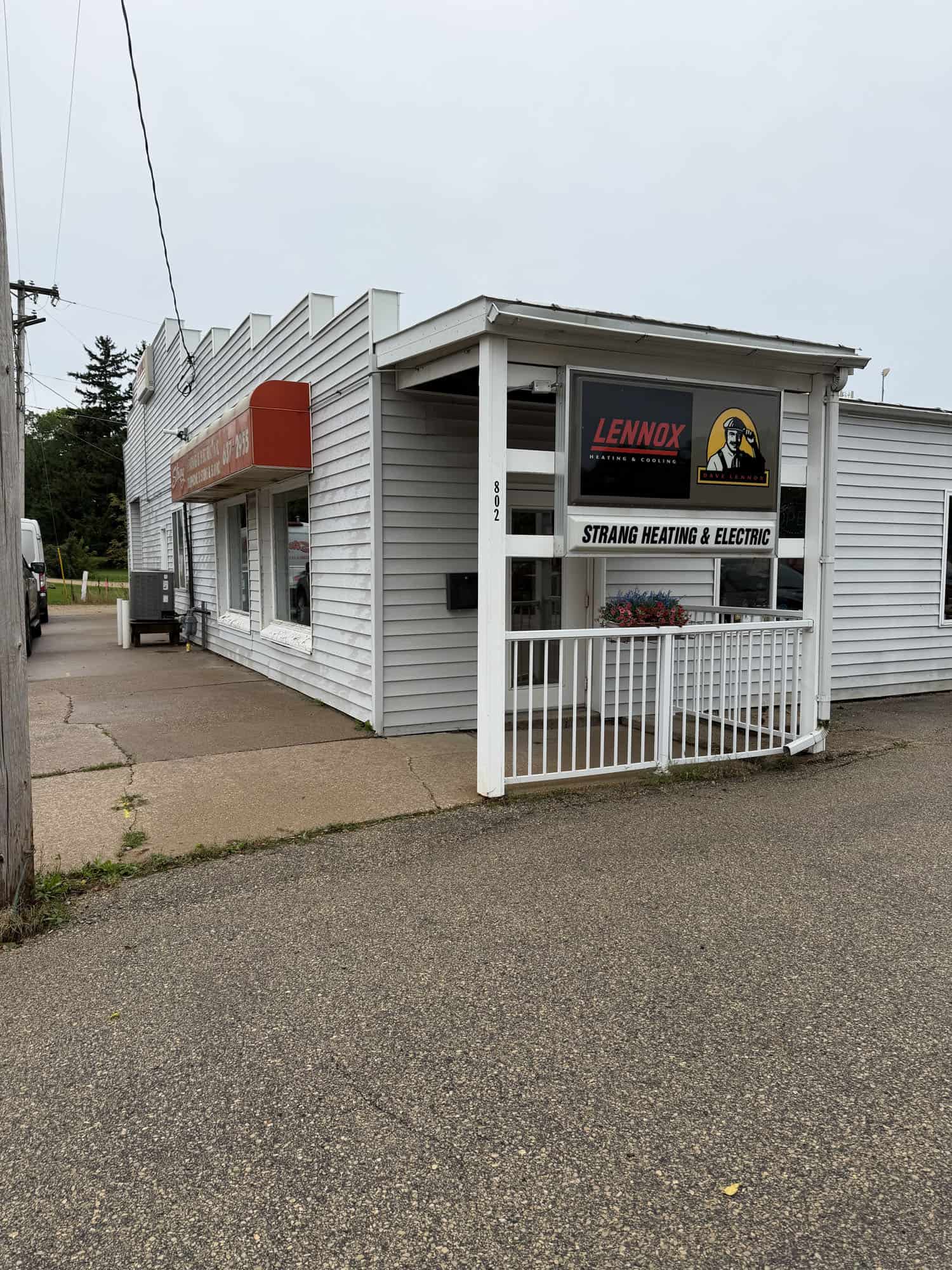 A small white building houses Strang Heating & Electric with a Lennox sign above the entrance. A red awning runs along the side, and a white railing borders the front porch. The street and sidewalk are empty.
